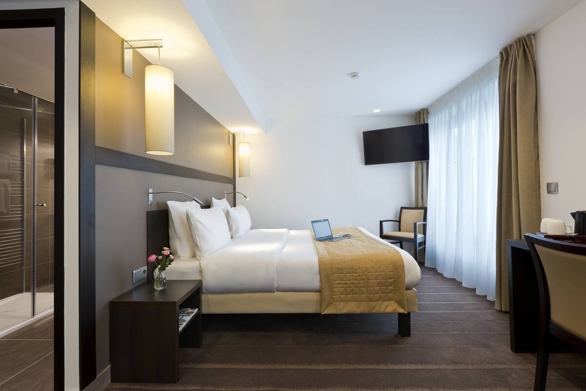 A hotel room with a bed, bedside table, lamp, TV, and a desk with a chair.