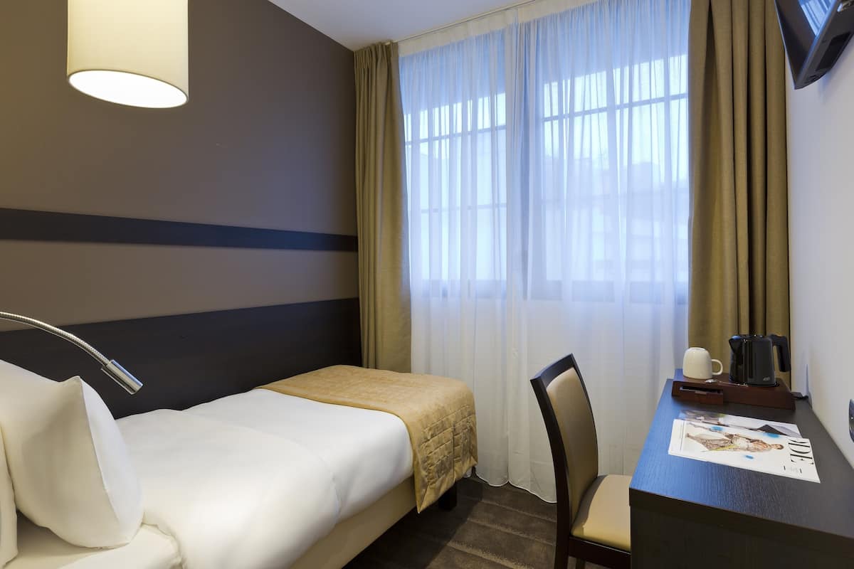 A hotel room with a bed, a desk with a coffee maker, a chair, and a window with curtains.