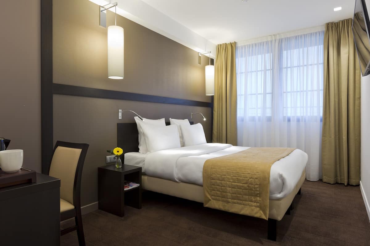 A hotel room with a bed, bedside table, chair, and a desk with a lamp.