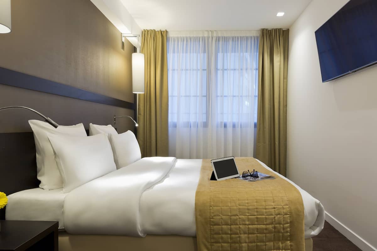 A hotel room with a bed, a bedside table, a lamp, a TV, and a window with curtains.