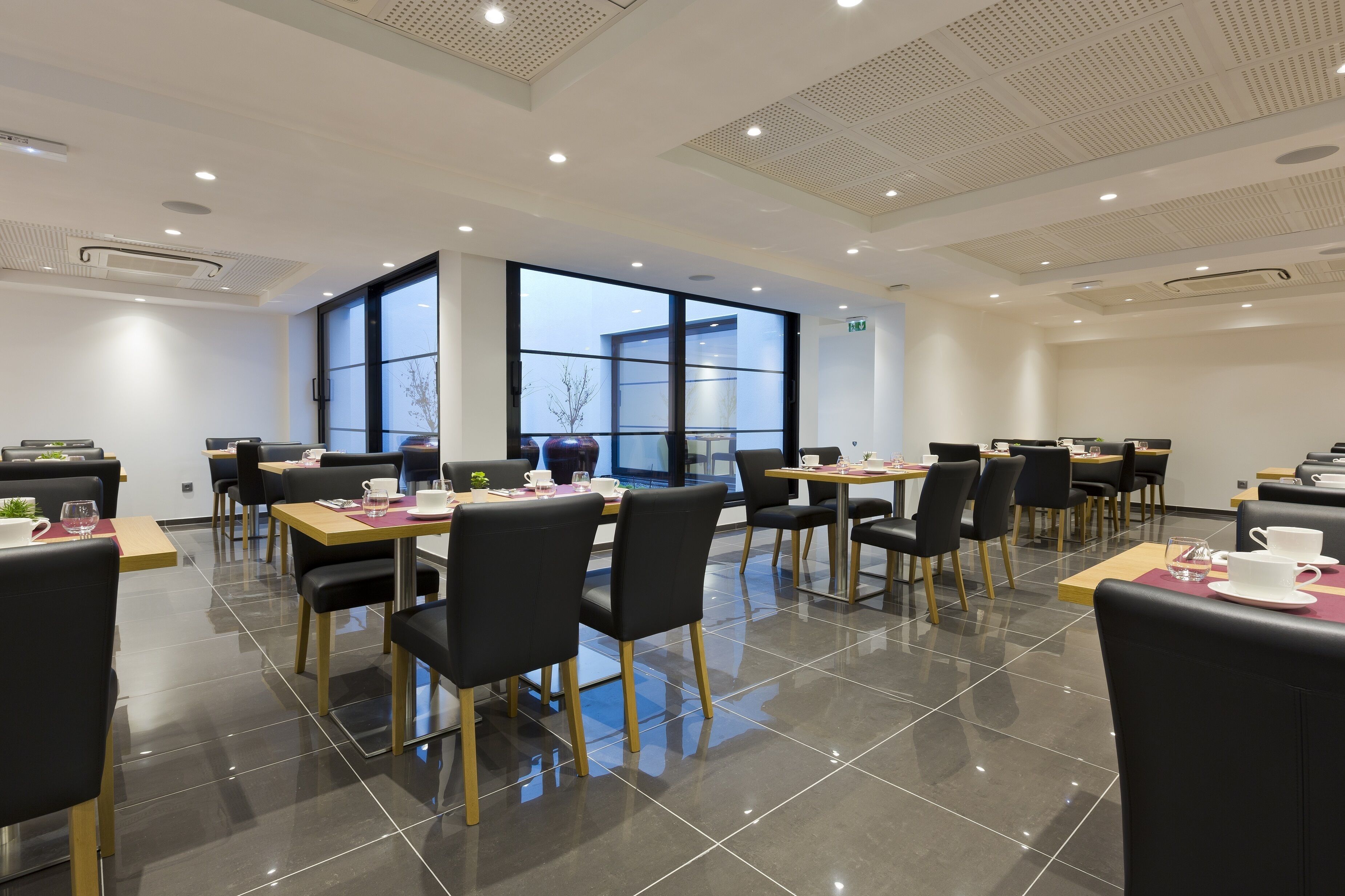 Un comedor moderno con mesas y sillas, ventanas amplias y un piso de baldosas limpias.