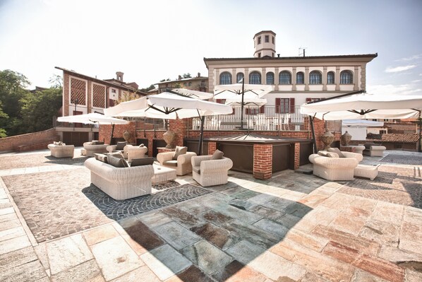 An outdoor patio with wicker furniture and umbrellas, a brick fireplace, and a large building in the background. - Relais Villa Prato (Mombaruzzo)