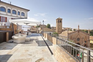 Terraço no telhado com soalho de pedra, mobiliário de exterior e vista para edifícios históricos, incluindo uma torre de igreja.