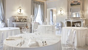 Un comedor lujoso con mesas redondas dispuestas para una comida, sillas elegantes y una chimenea al fondo.