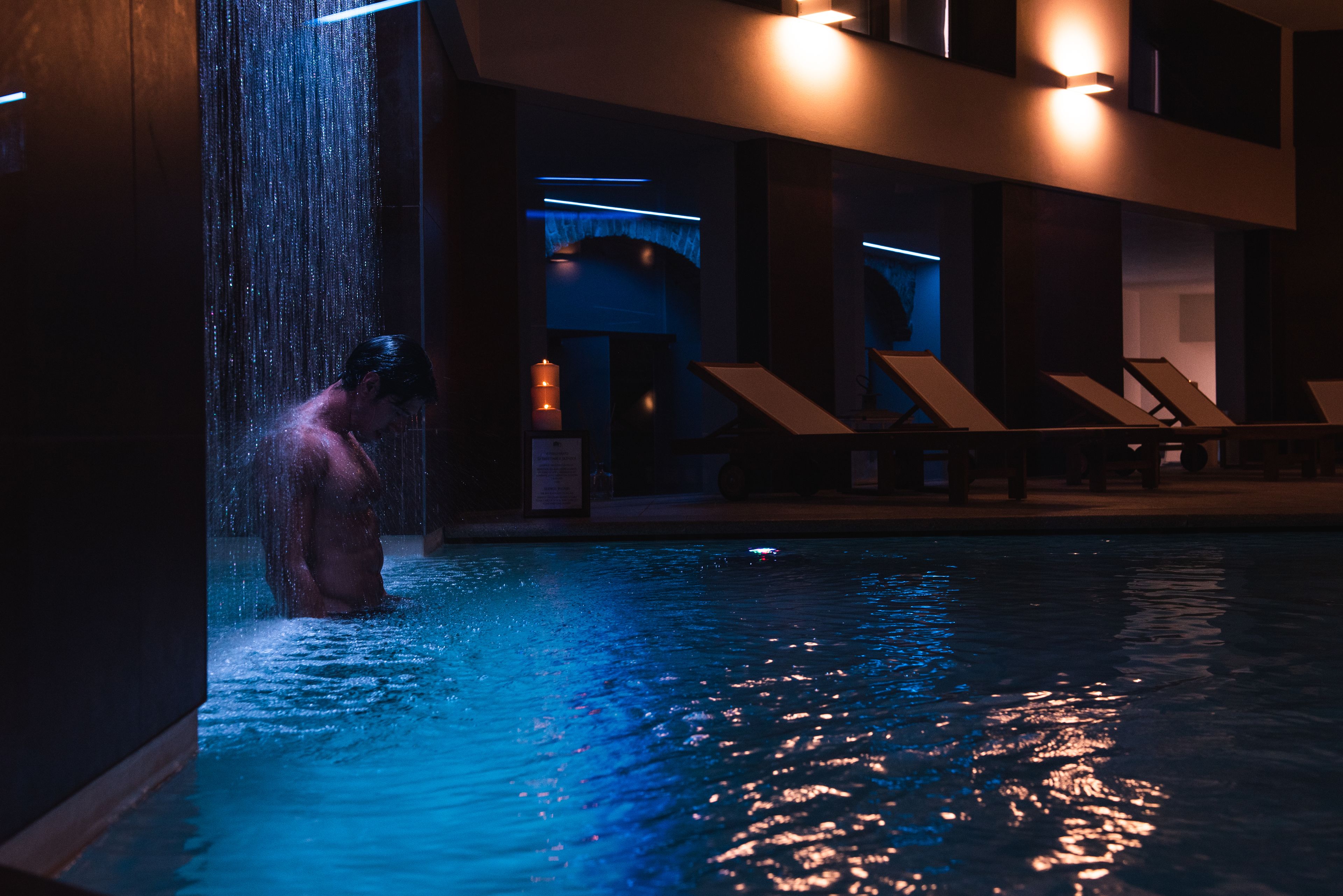 Ein Mann in einem Pool mit Wasserstrahlen, umgeben von Liegestühlen und moderner Architektur.