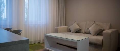 Una sala de estar moderna con una mesa de centro de vidrio, un sofá color beige con almohadones y una ventana con cortinas translúcidas.