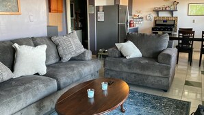 A living room with two grey sofas, a wooden coffee table, and a kitchen area with a refrigerator, stove, and shelves.