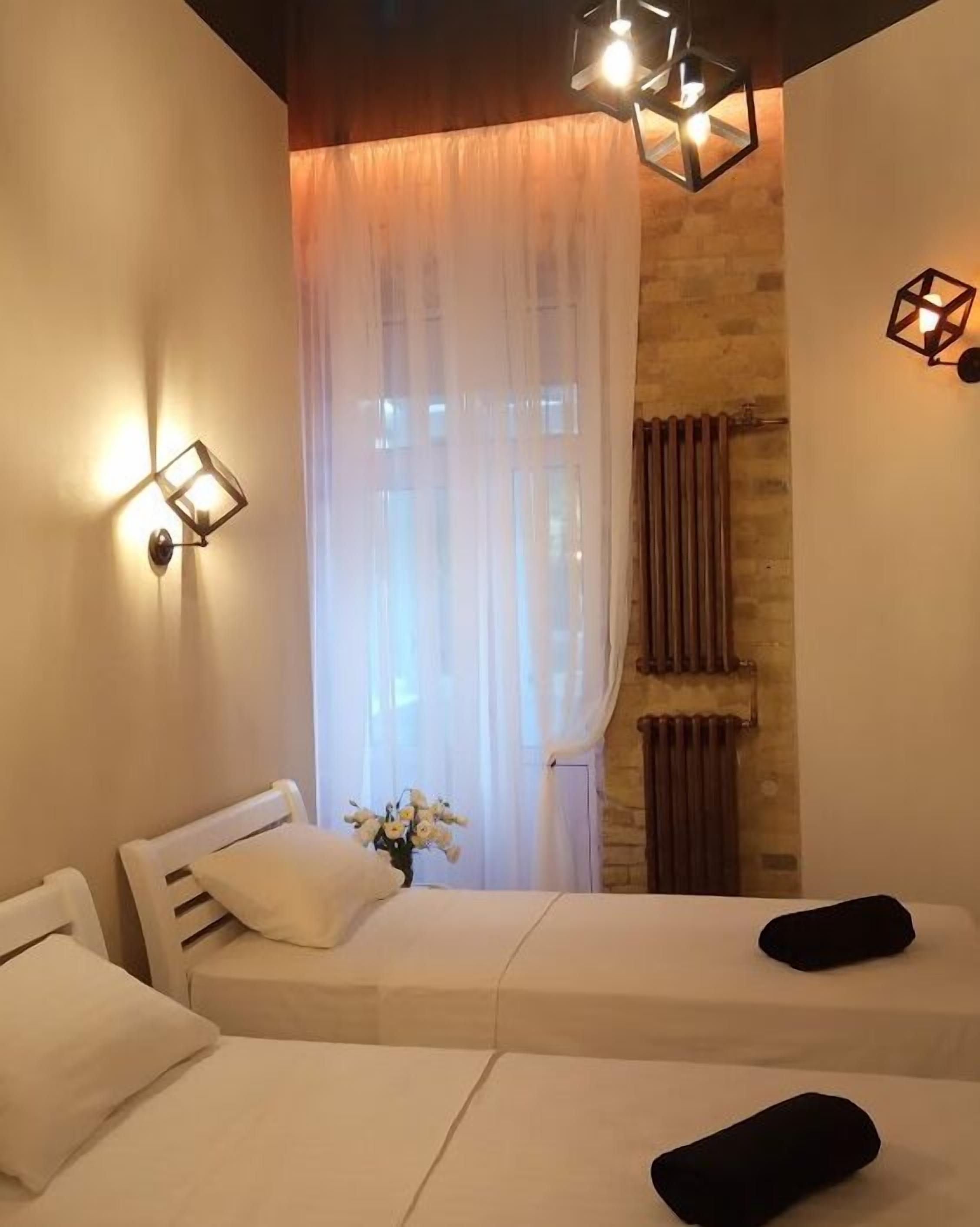 A room with two beds, a window with sheer curtains, a radiator, and wall-mounted lights.