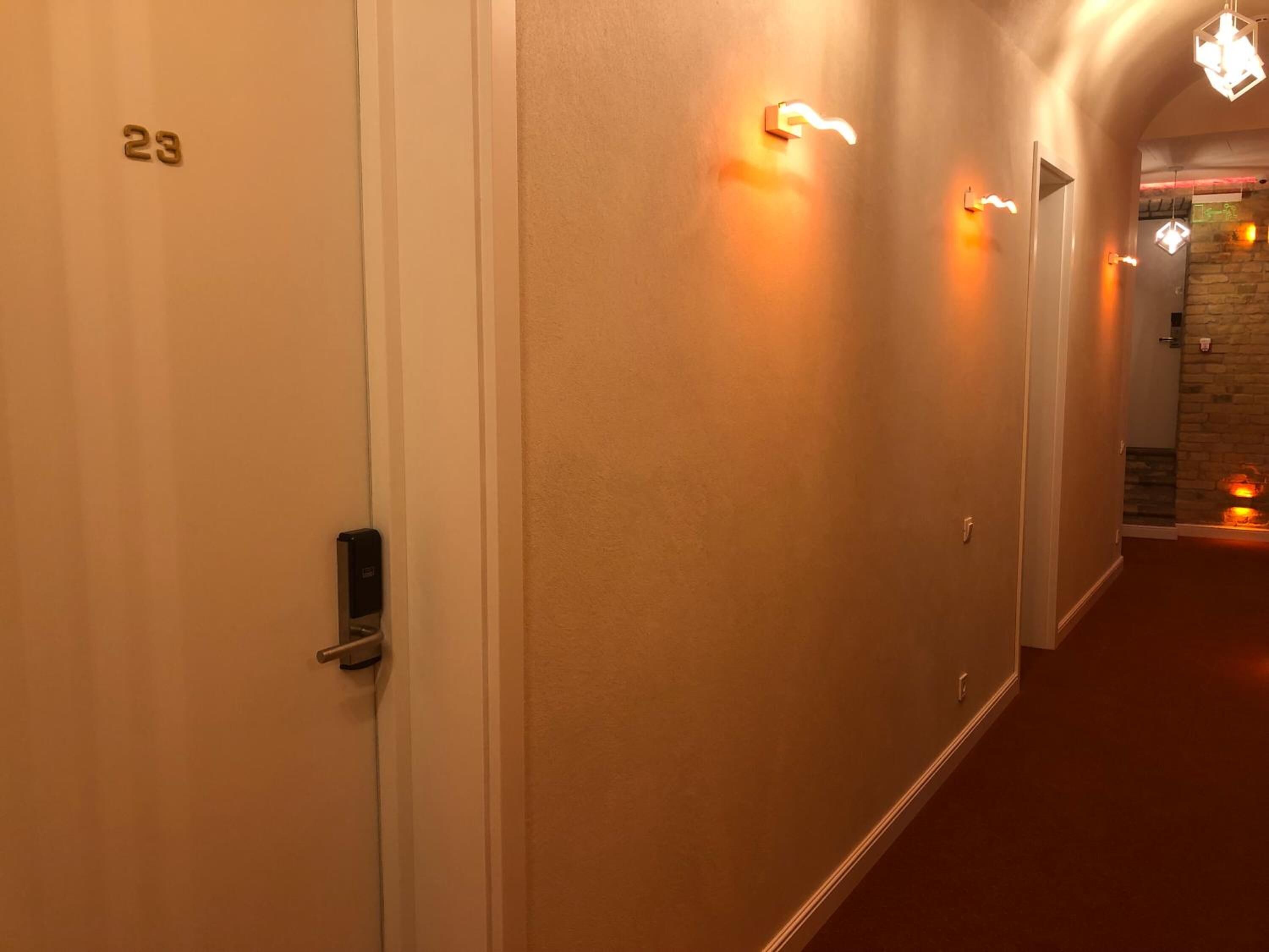 A hallway with a door labeled '23', a wall-mounted light fixture, and a stone wall in the background.
