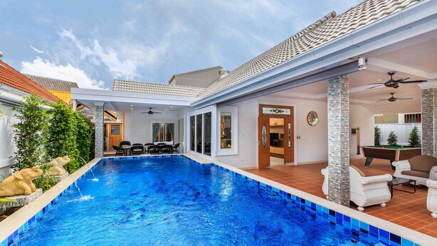 Una piscina con borde de azulejos, un porche cubierto con mobiliario exterior y vista del edificio contiguo.