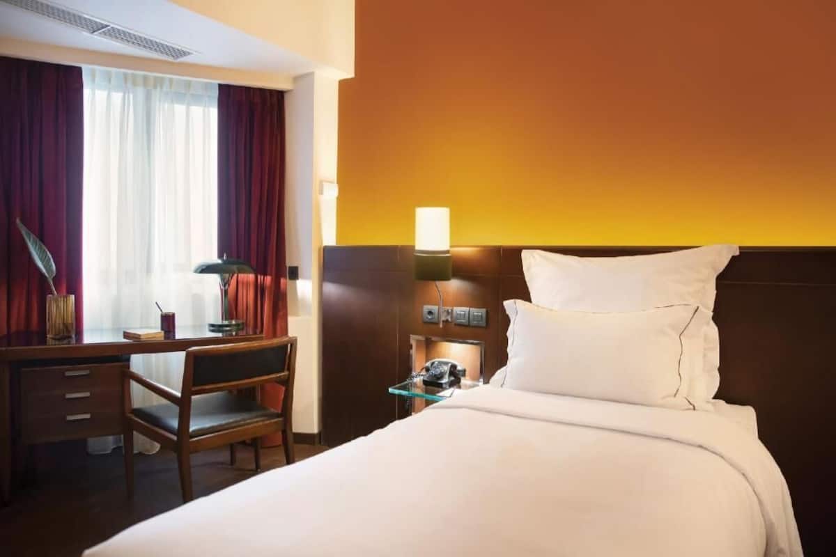 Quarto de hotel com cama, escrivaninha, cadeira, abajur e telefone.