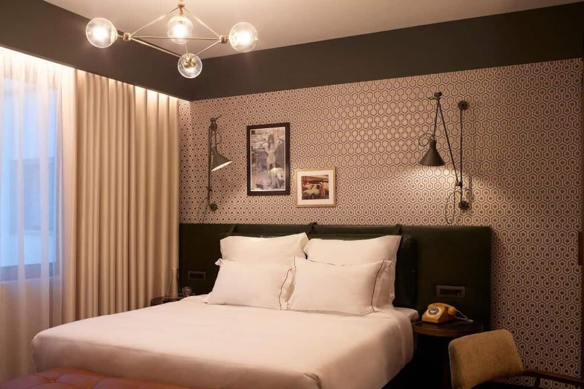 Quarto de hotel com uma cama grande, cabeceira, mesa de cabeceira com telefone, uma cadeira e janela com cortinas.