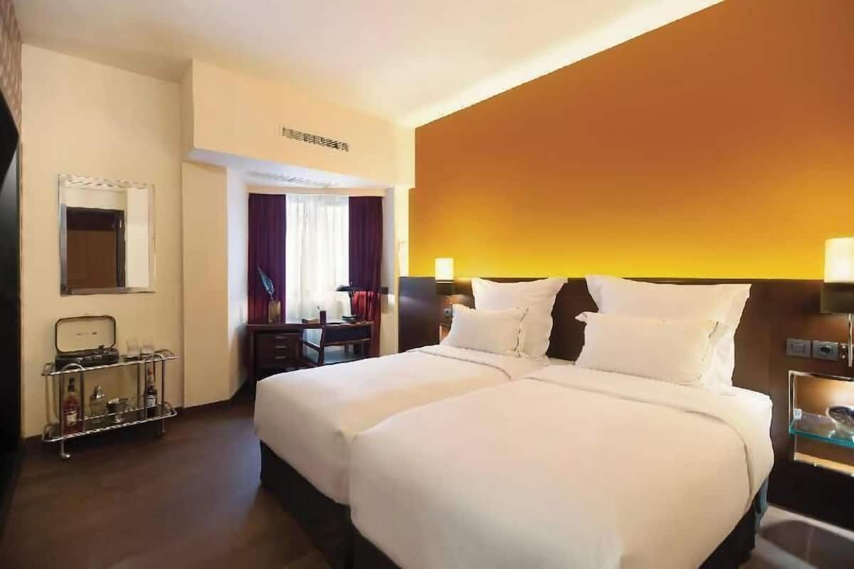 Quarto de hotel com duas camas, uma escrivaninha, uma cadeira, um minibar e uma televisão.