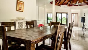 Uma sala de jantar espaçosa com uma grande mesa de madeira, seis cadeiras e um vaso com flores vermelhas. H á um quadro na parede e um alto-falante em um tripé ao fundo.