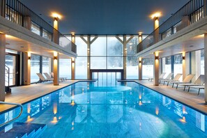 A large indoor swimming pool with a wooden deck, sun loungers, and a view of the snowy landscape through large windows. - Hotel Pustertalerhof (Chienes)