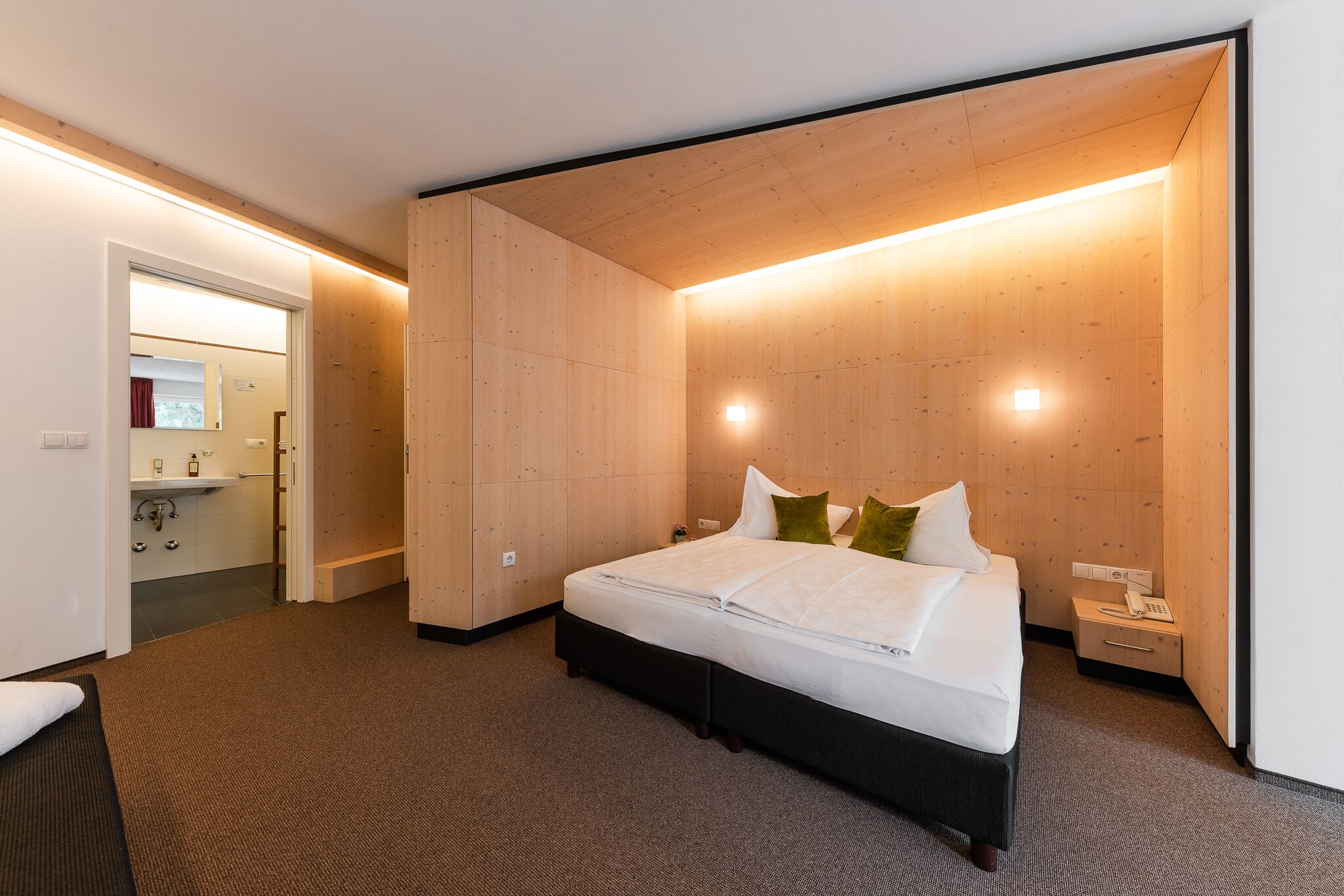 Una habitación de hotel moderna con una cama grande, paneles de madera, una mesita de noche con teléfono y un baño visible.