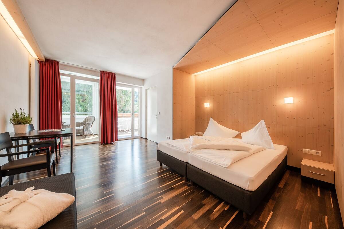 Une chambre d’hôtel moderne dotée d’un grand lit, d’un plancher en bois, d’un coin repas avec une table et des chaises, et offrant une vue à travers une porte vitrée.