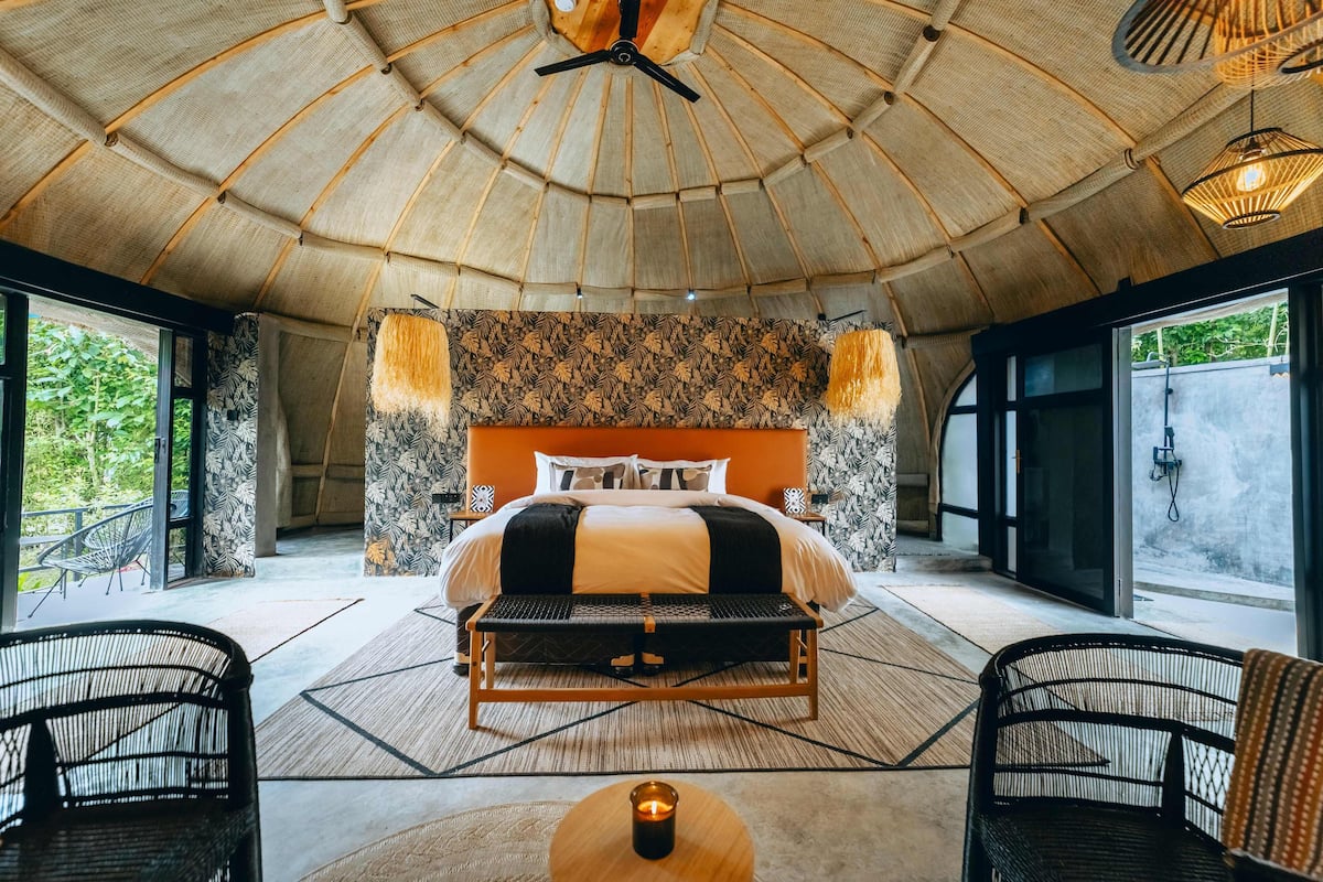 A spacious room with a large bed, two side tables, a bench, and a view of greenery outside.