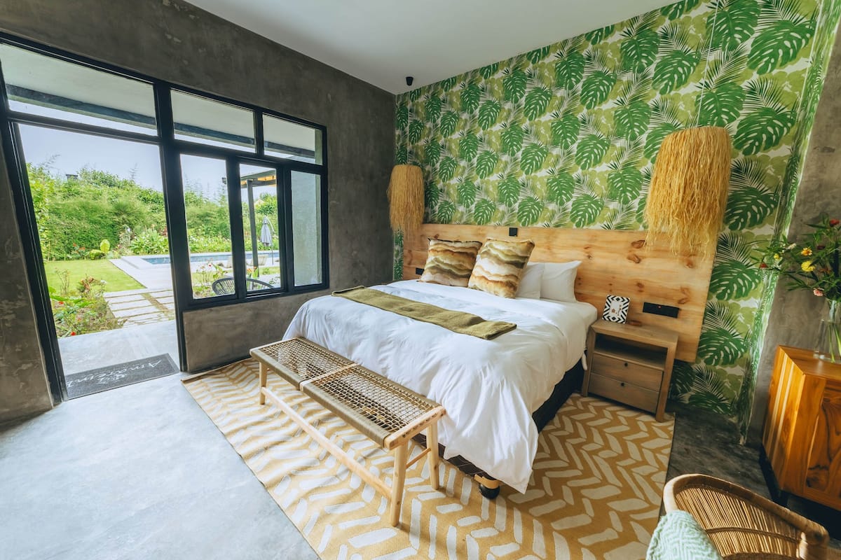 A modern bedroom with a large bed, wooden headboard, and a view of a garden through large windows.