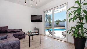 A modern living room with a sofa, a coffee table, and a TV. There's a sliding glass door leading to a pool area with a view of the outdoors. - Luxurious home in Betina (Betina)