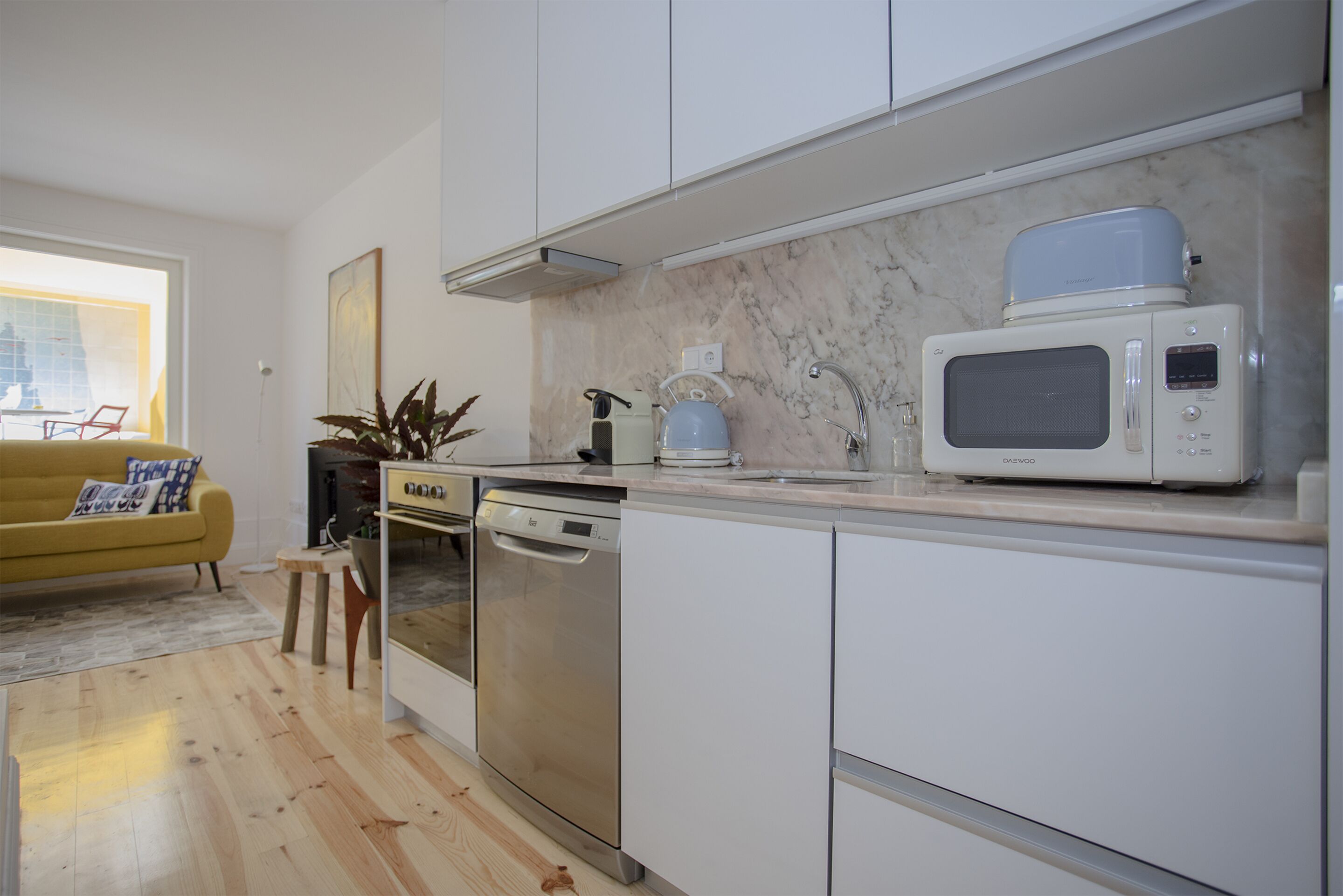 Une cuisine moderne avec des meubles blancs, un lave-vaisselle, un four à micro-ondes et une machine à café. Le sol est en bois et le salon adjacent est doté d’un canapé jaune.