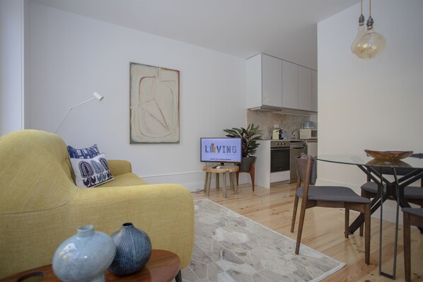 Un salon moderne avec un canapé, une table basse ornée de vases, un coin repas avec des chaises, une cuisine équipée de placards et un téléviseur affichant le mot « VIVRE ».