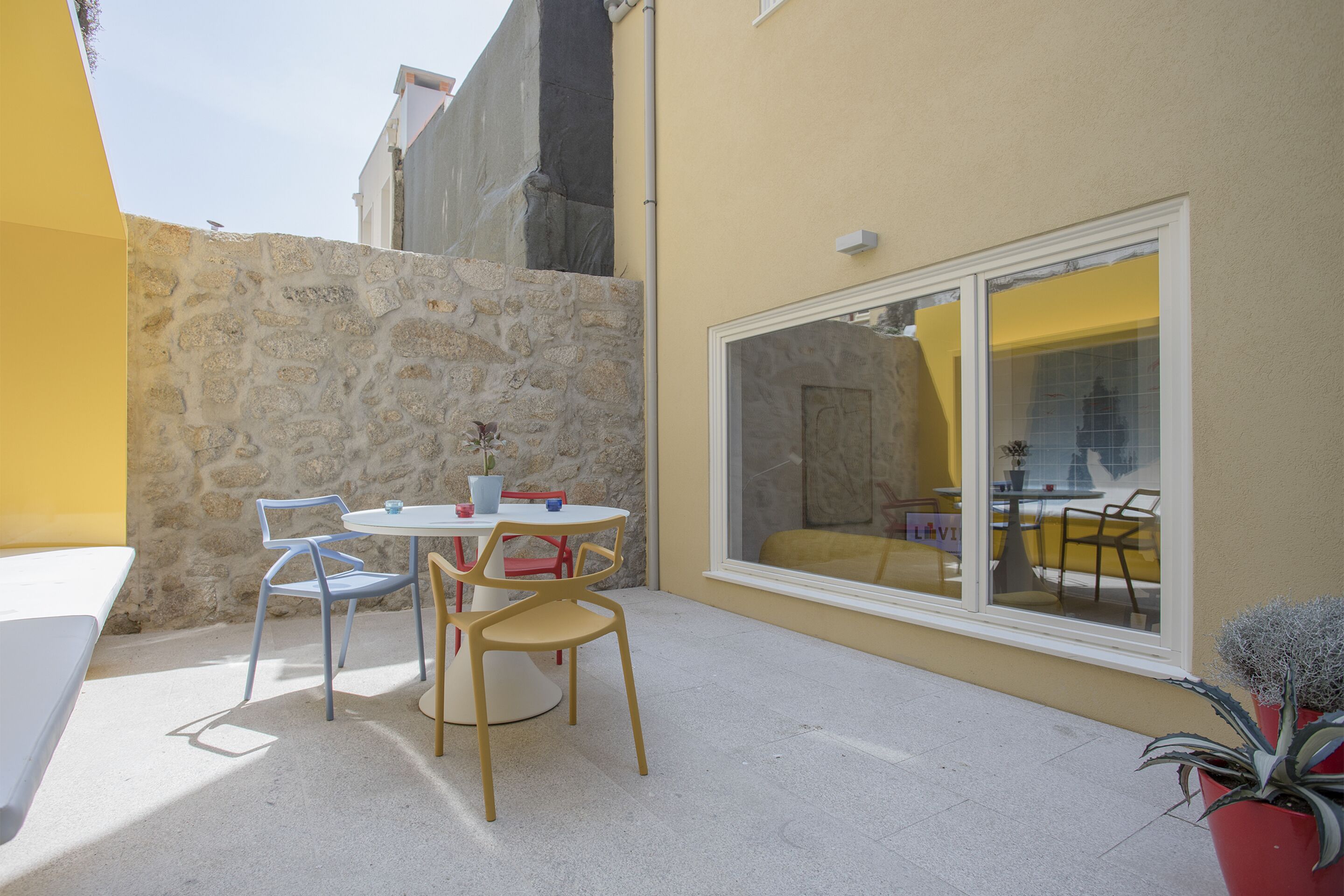 Une terrasse extérieure avec une table ronde et des chaises, un mur en pierre et une fenêtre dont l’intérieur est jaune.