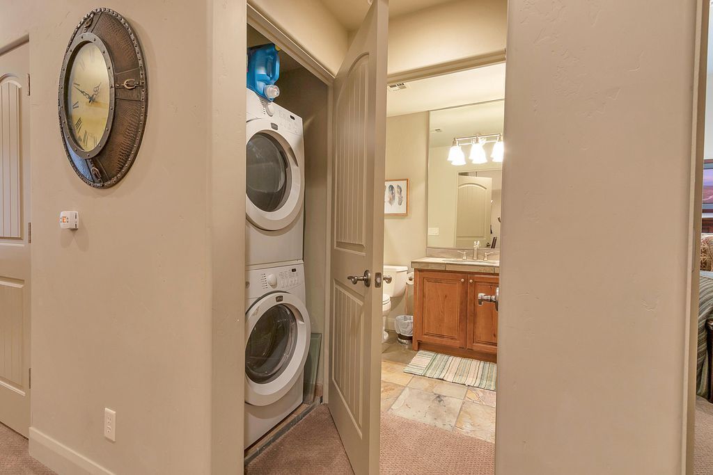 Une buanderie équipée de deux machines à laver et deux sèche-linge superposés, une horloge au mur et une porte donnant sur une salle de bain avec un lavabo et un miroir.