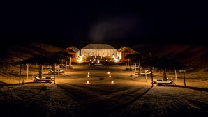 A well-lit resort entrance with a large building, palm trees, and lounge chairs. - Erg Chebbi Camp (Taouz)