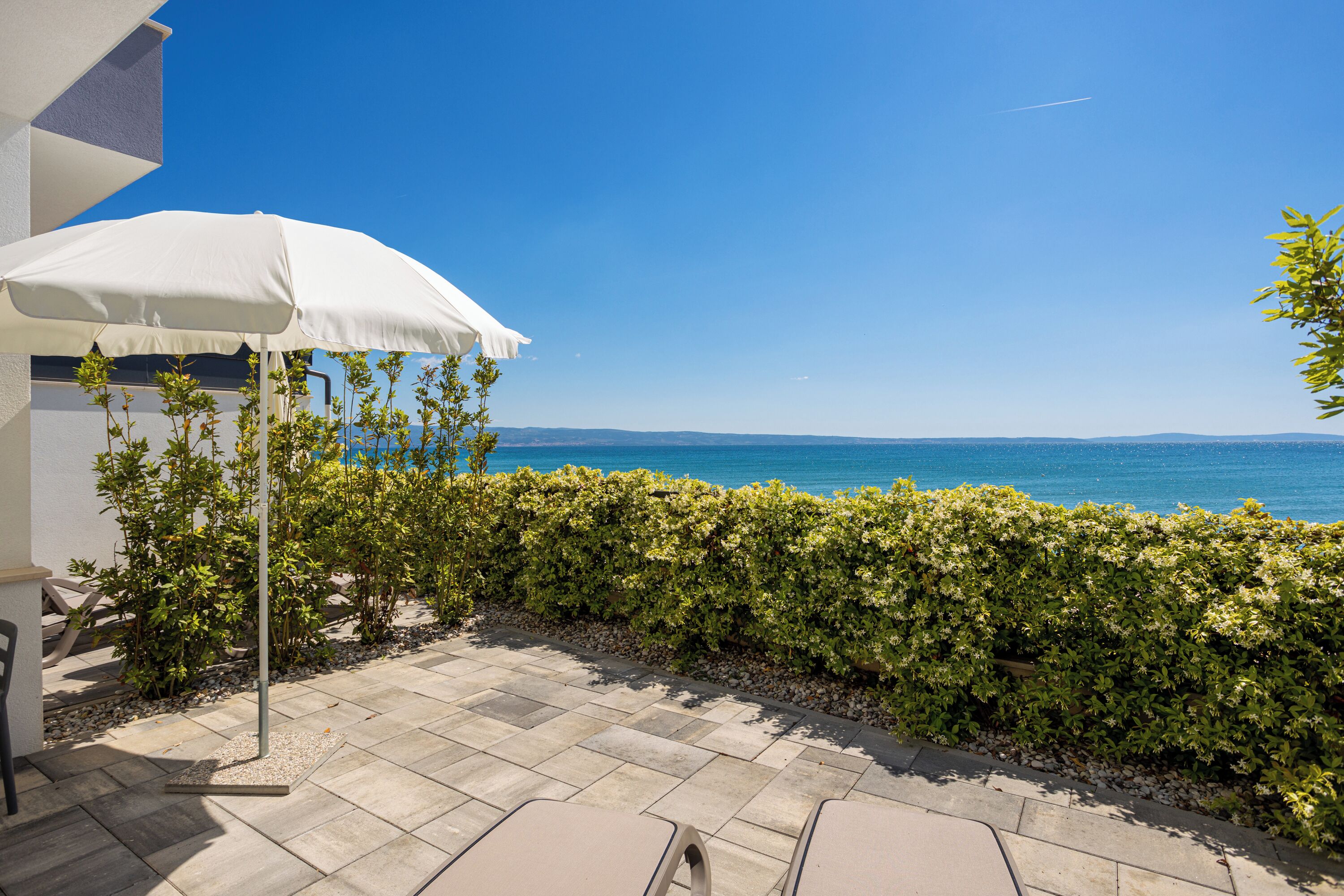 Un patio con un ombrellone bianco, vista mare e un siepe.