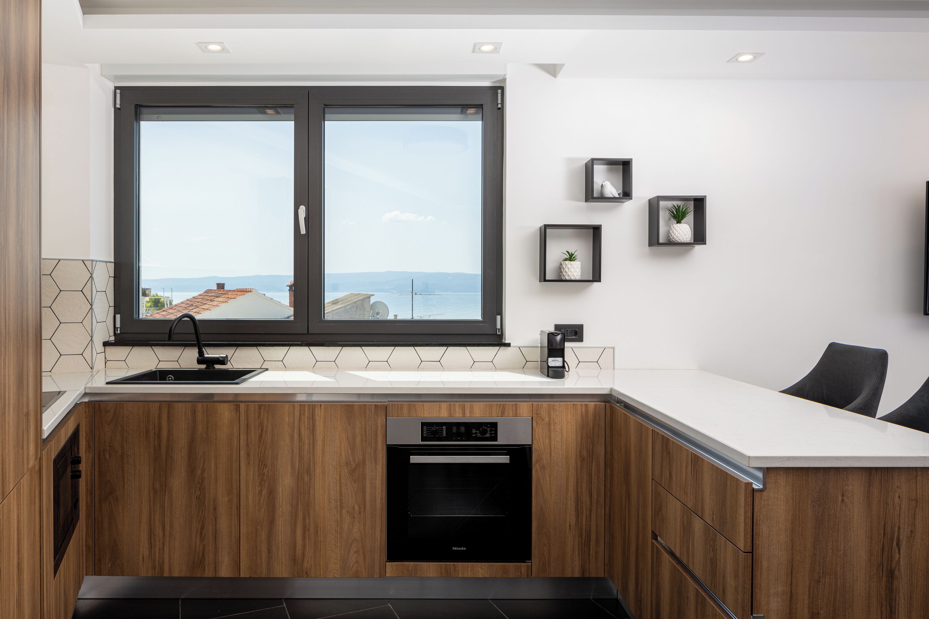 A modern kitchen with wooden cabinets, a built-in oven, a large window with a view, and a small countertop with a sink.