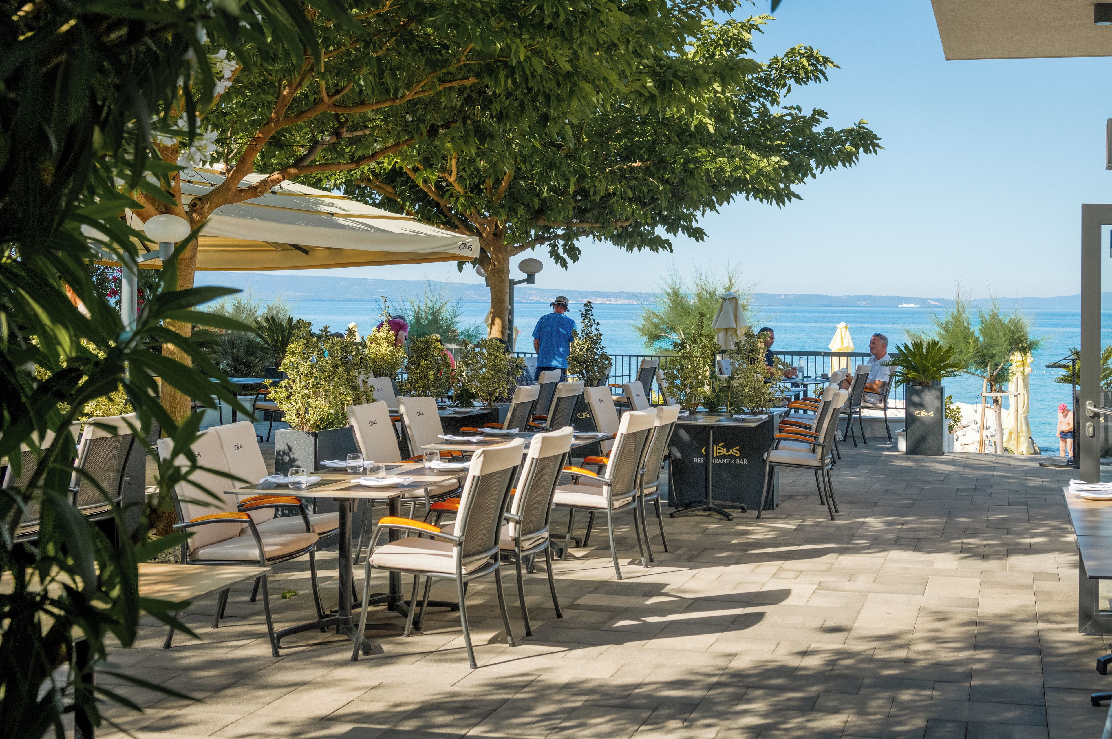 Zona pranzo all'aperto con tavoli e sedie, affacciata su una distesa d'acqua.