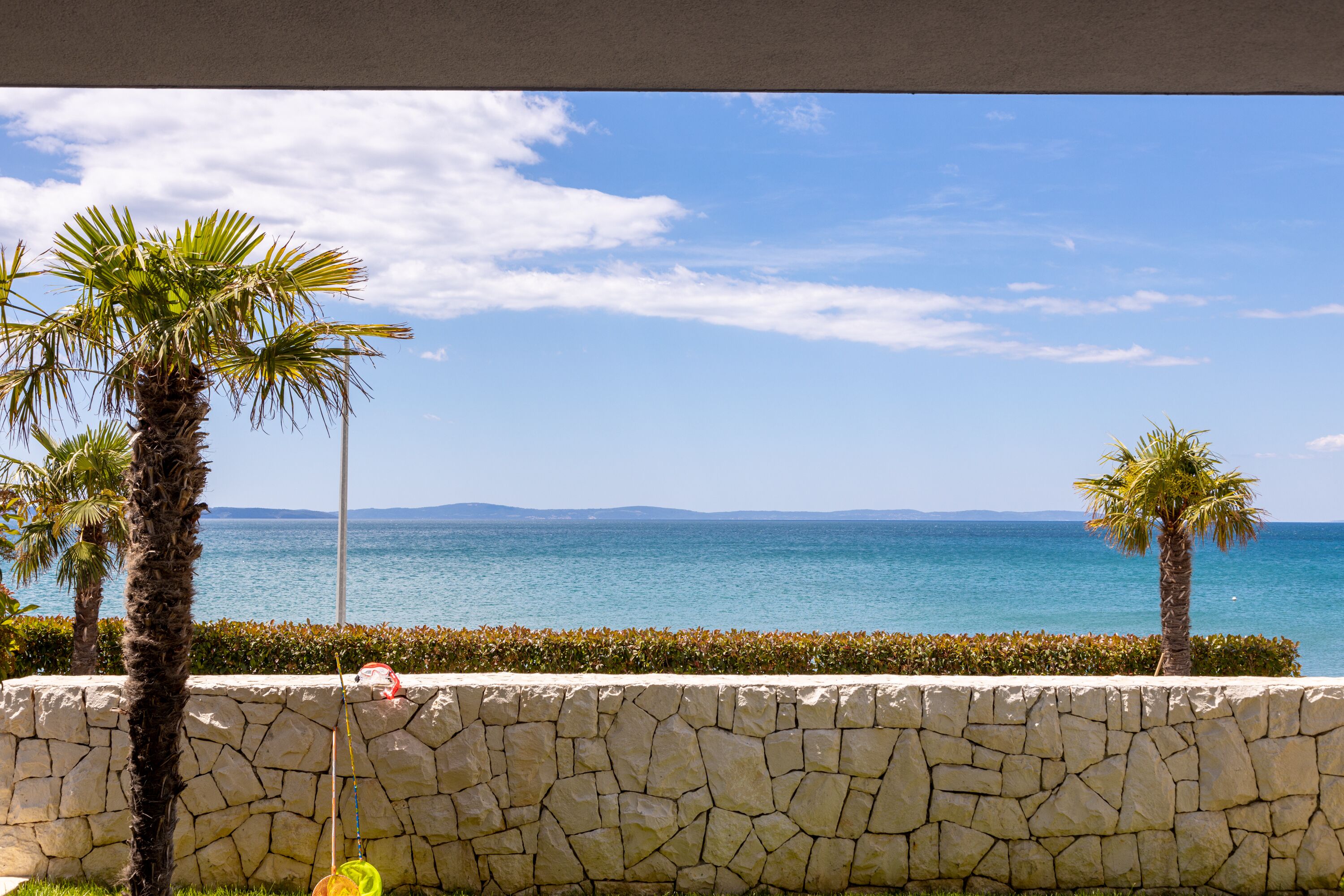 Vista sulla costa con un muro di pietra, palme e un cielo azzurro limpido.