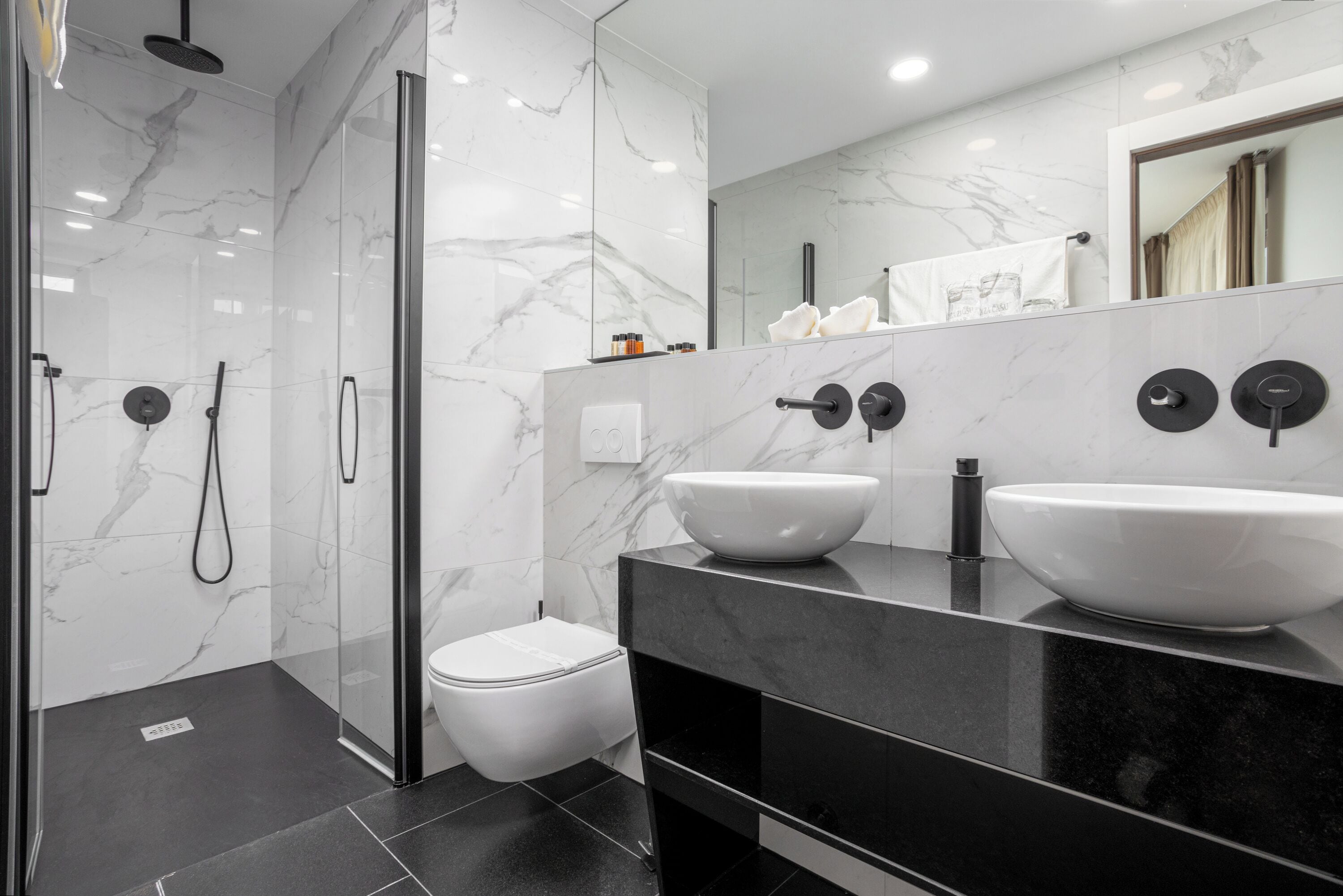 Een moderne badkamer met een douchecabine van glas, een wit toilet en twee wasbakken op een zwart aanrecht.