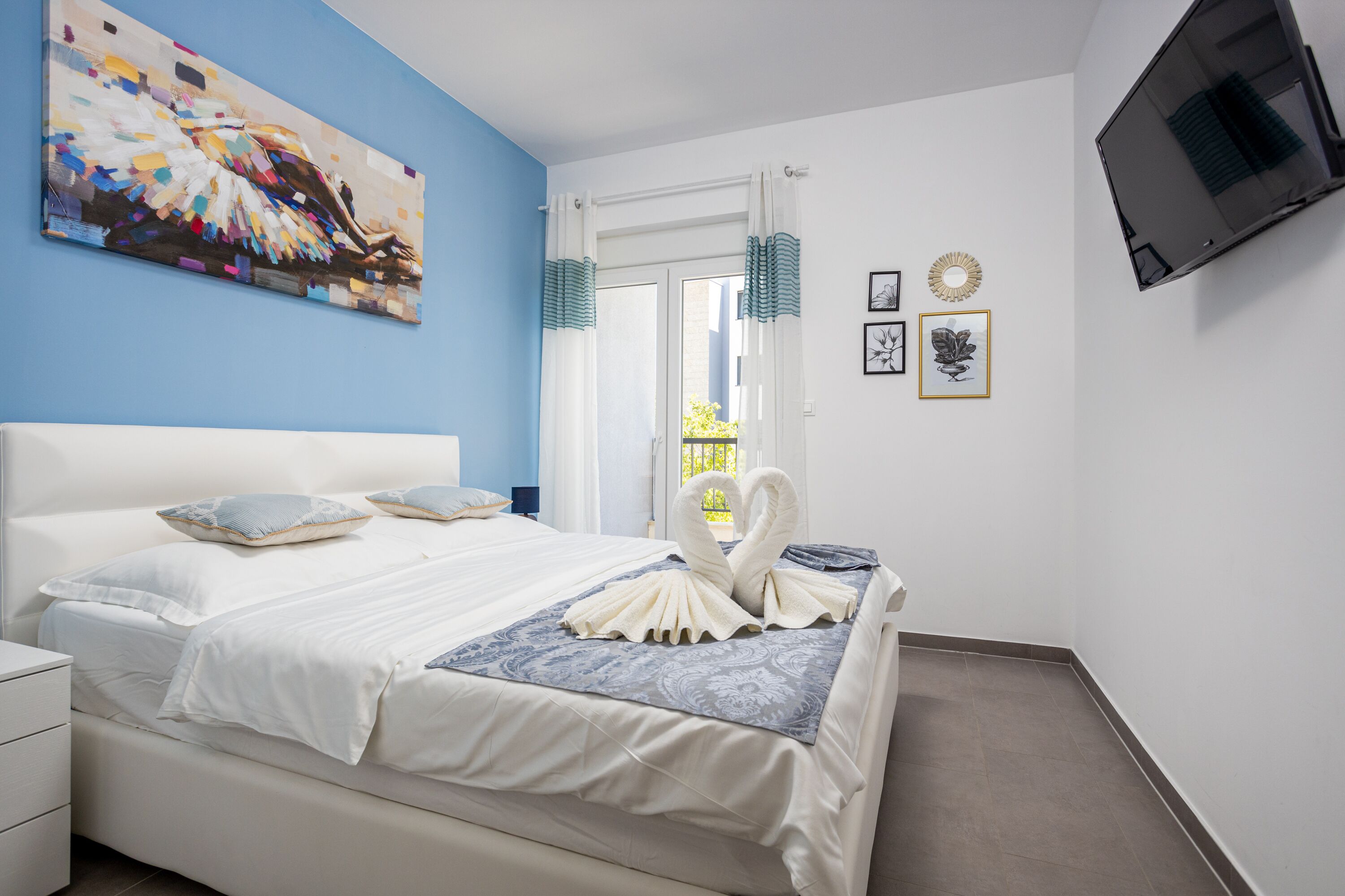 Ein ordentlich bezogenes Bett mit einer dekorativen Schwanen-Handtuch-Arrangement, ein großes abstraktes Gemälde an der Wand und ein an der Wand befestigtes Flachbildfernsehen.
