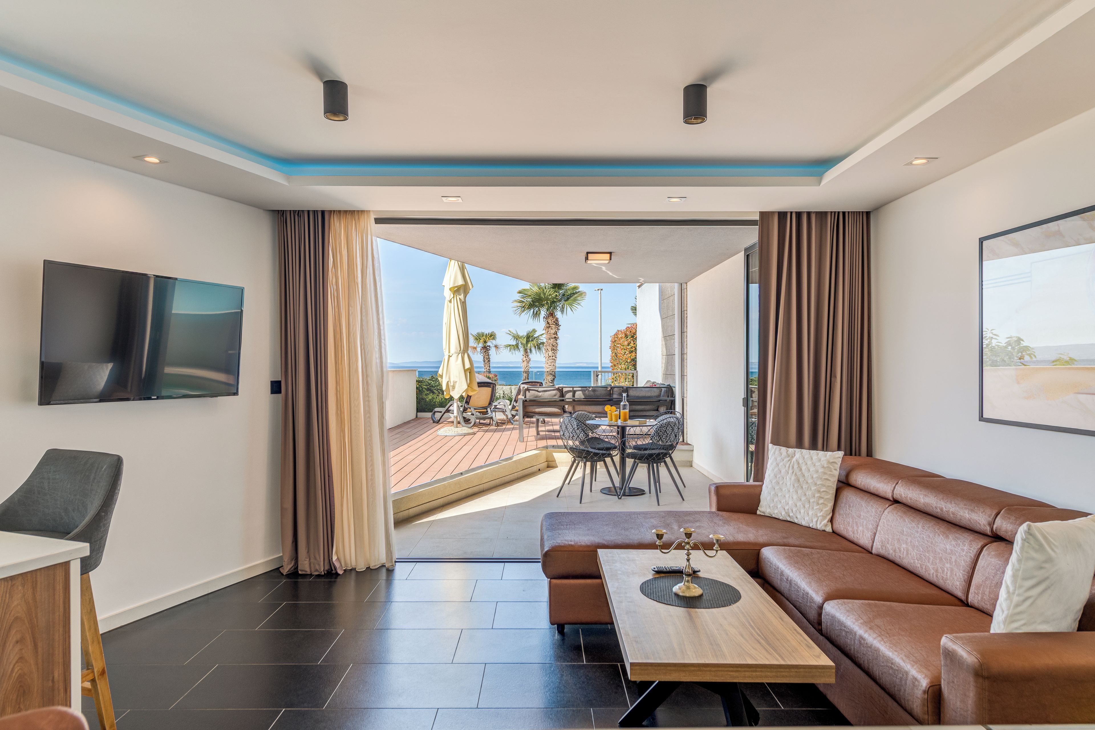 A modern hotel room with a large sofa, a flat-screen TV, a dining table, and a view of the sea.
