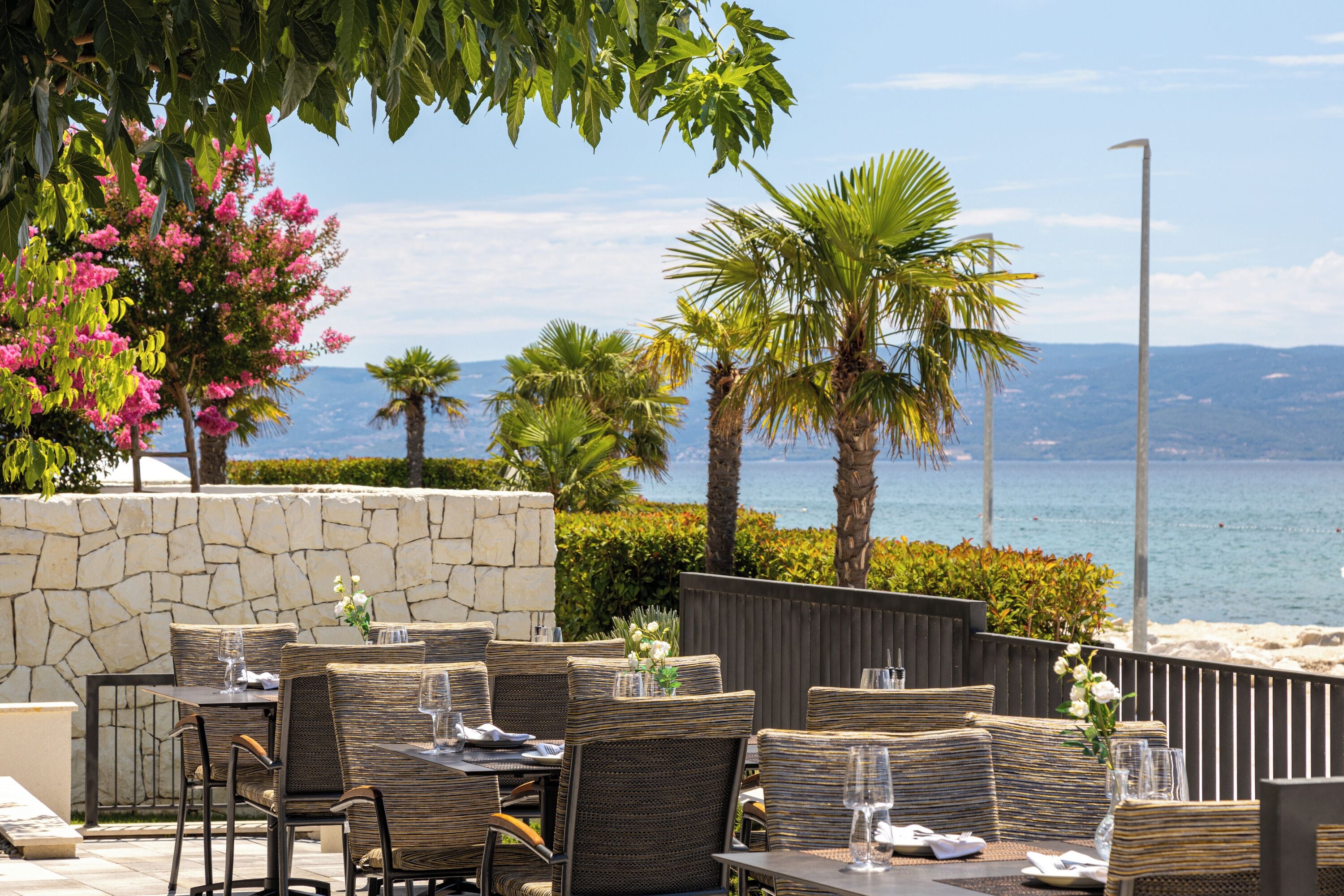 Un comedor al aire libre con mobiliario de mimbre, una pared de piedra y vistas al mar.