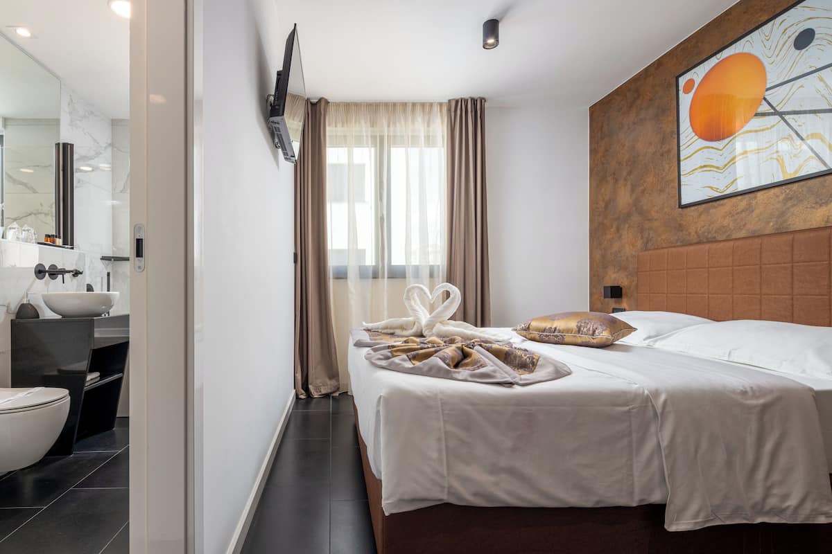 A hotel room with a bed, a bathroom, a large window with curtains, and a wall-mounted TV.