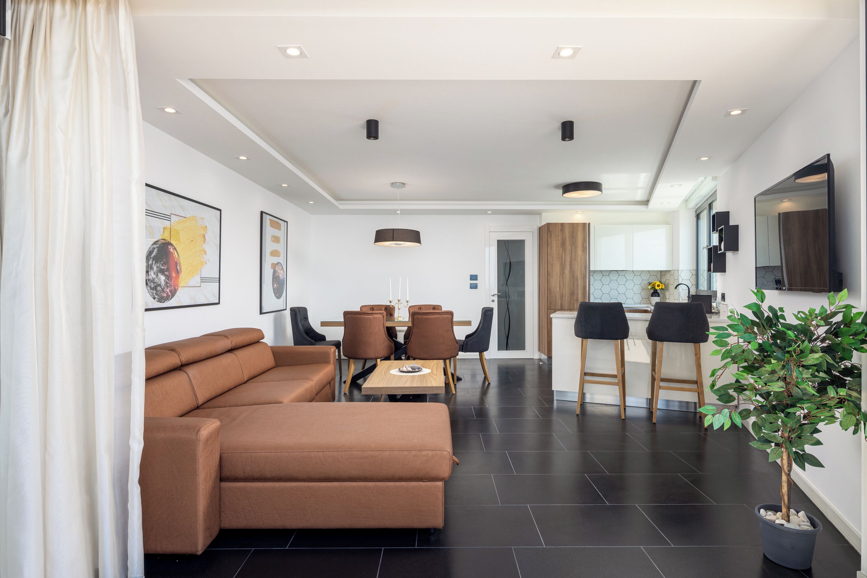 A modern living room with a brown sectional sofa, a dining area with a wooden table and chairs, and a kitchen area with bar stools.