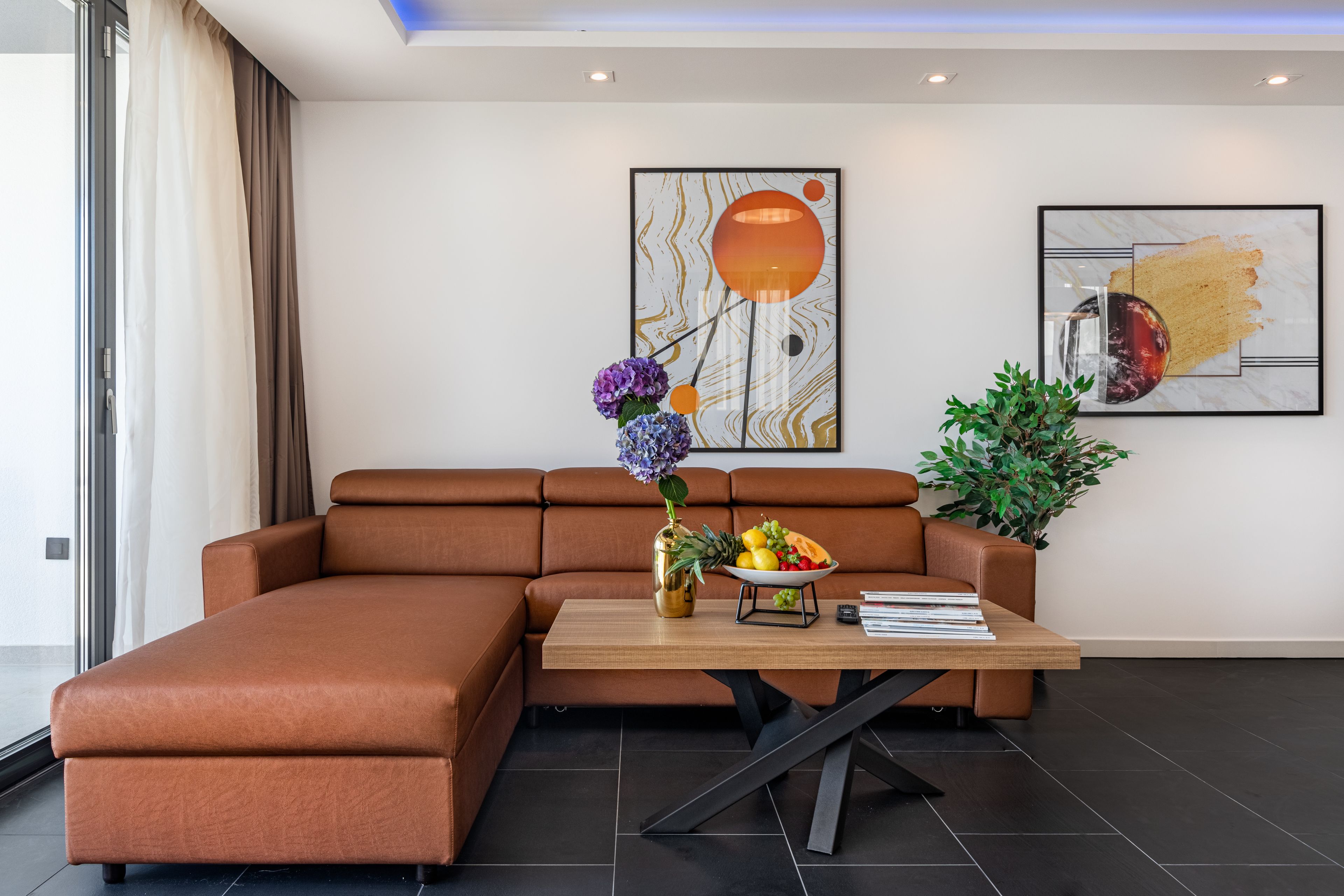 Un salon moderne avec un canapé d’angle marron, une table basse en bois sur laquelle se trouvent un panier de fruits et des livres, et deux tableaux abstraits accrochés au mur.