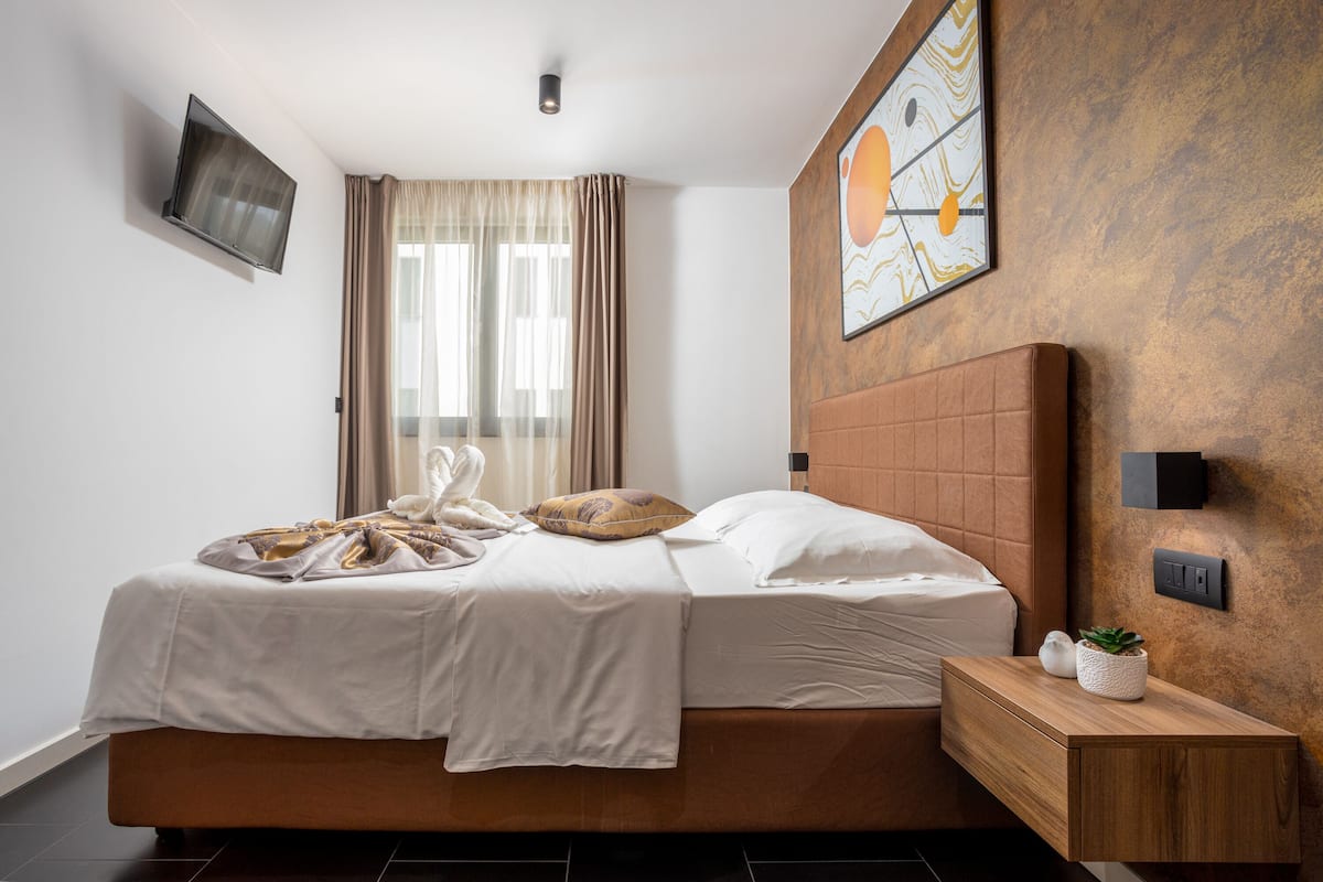 A modern bedroom with a large bed, a flat-screen TV mounted on the wall, a bedside table with a plant, and a large window with curtains.