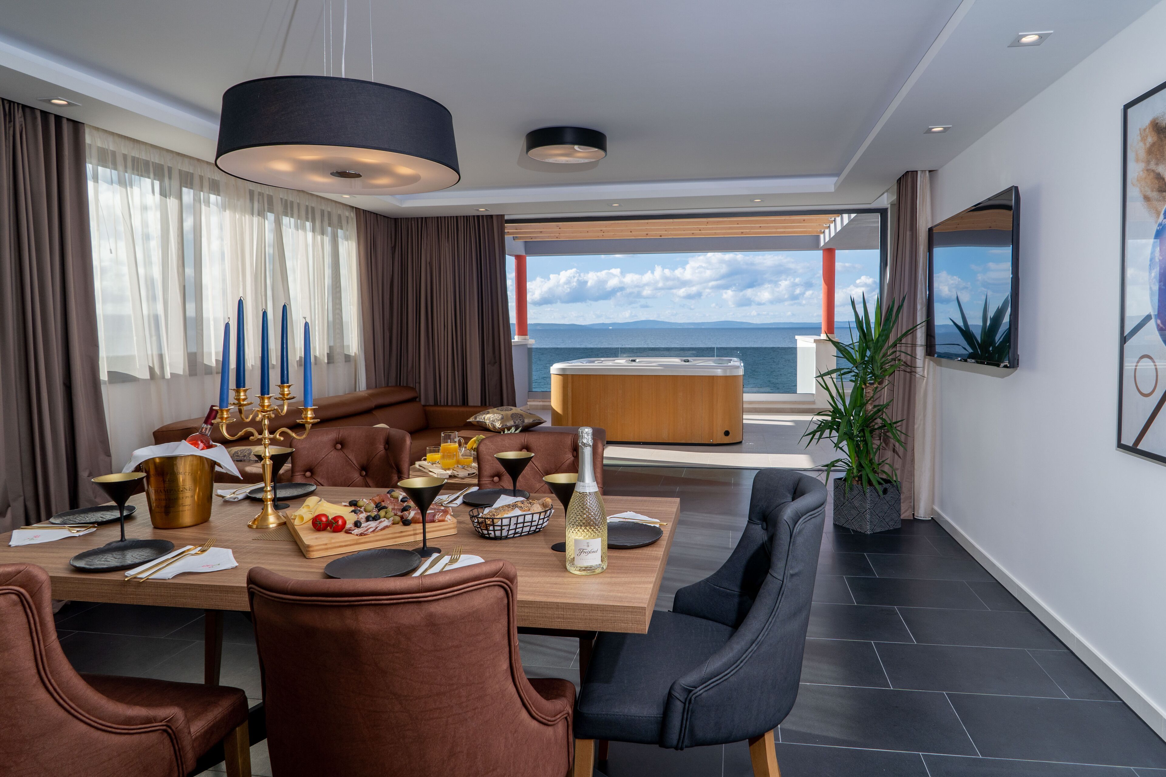 A modern dining area with a wooden table set for a meal, a large window offering a view of the sea, and a cozy seating arrangement.