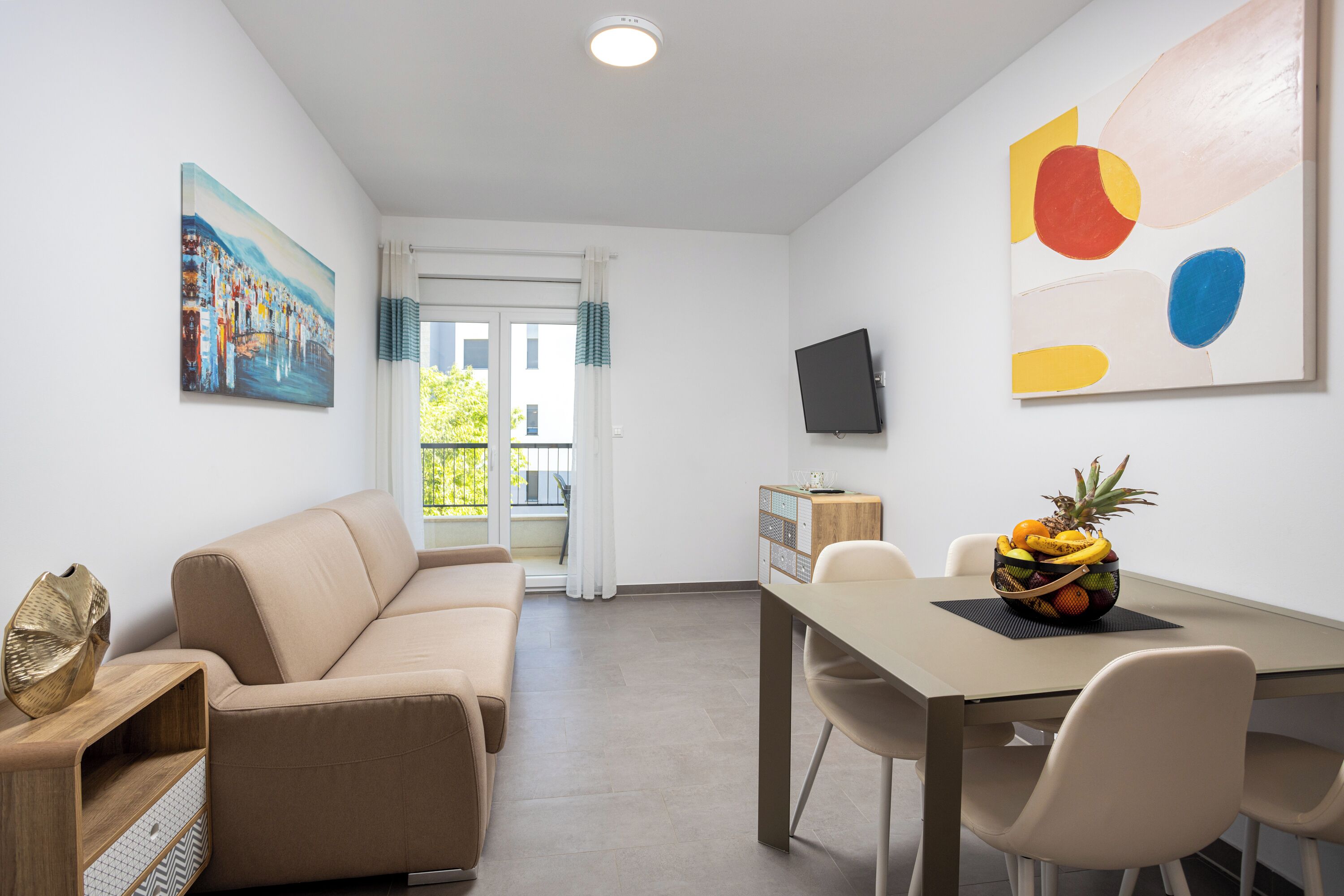 A modern living room with a sofa, a dining table with chairs, a fruit bowl, a TV on the wall, and a painting.