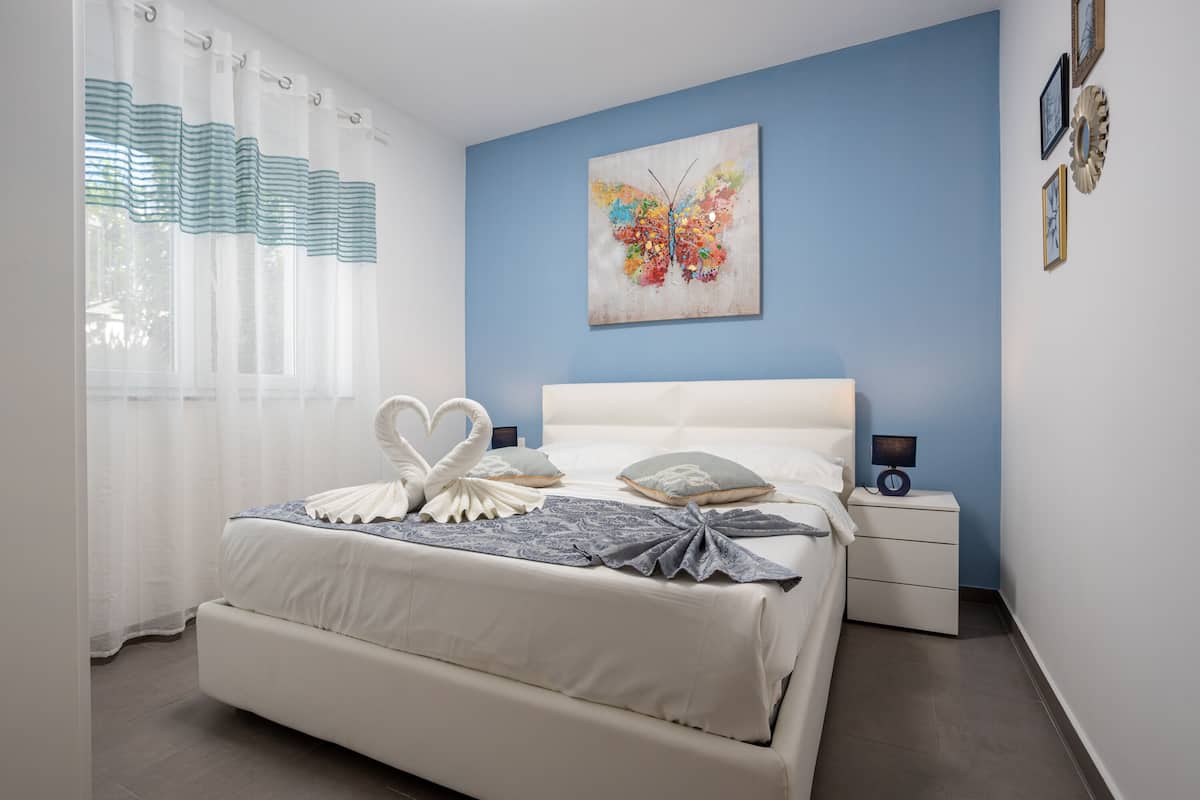 A bedroom with a bed, white curtains, a blue wall, and a butterfly painting.