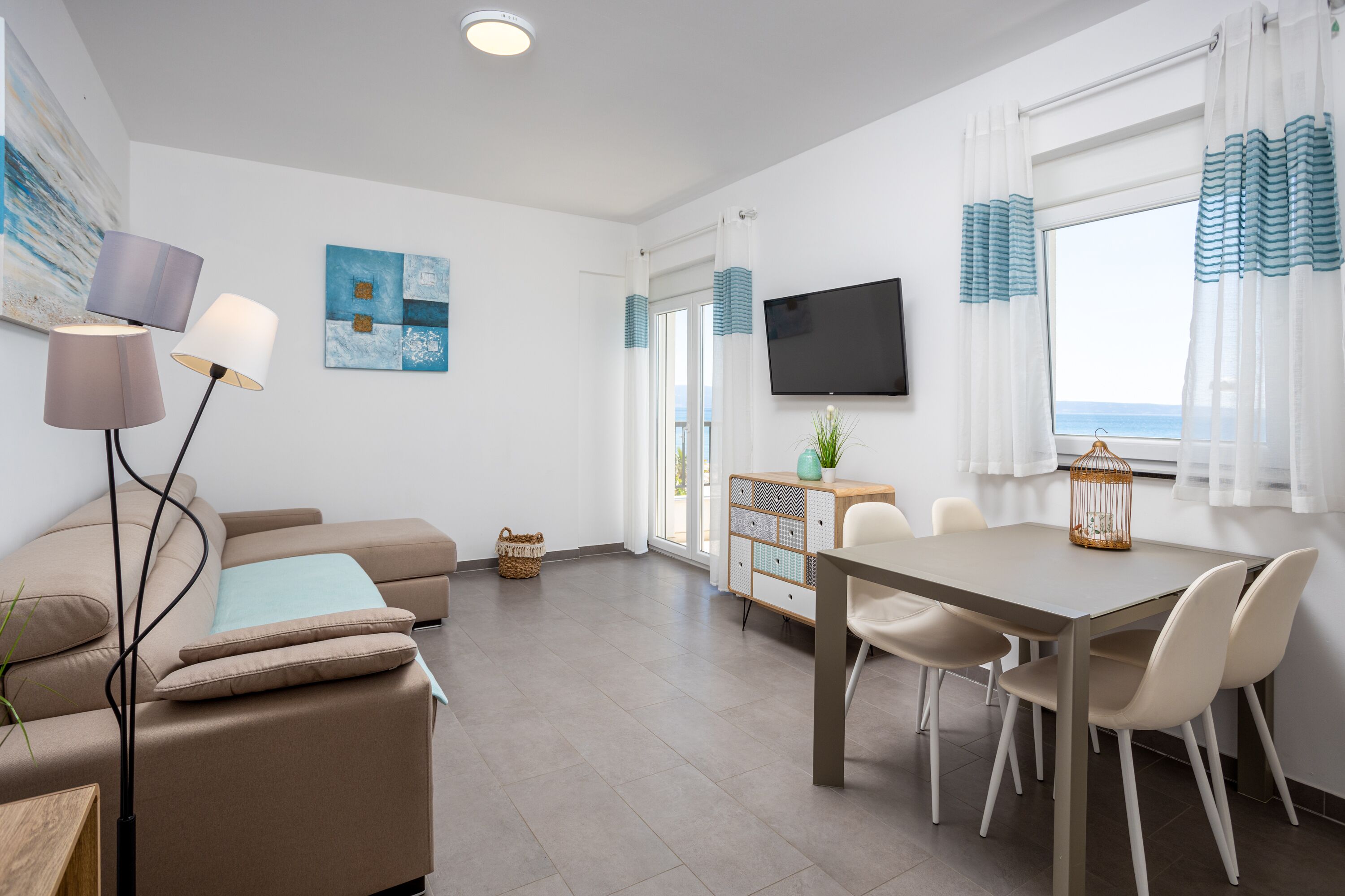 Una sala de estar moderna con sofá, una mesa de comedor con sillas, un televisor de pantalla plana y vista al mar a través de amplios ventanales.