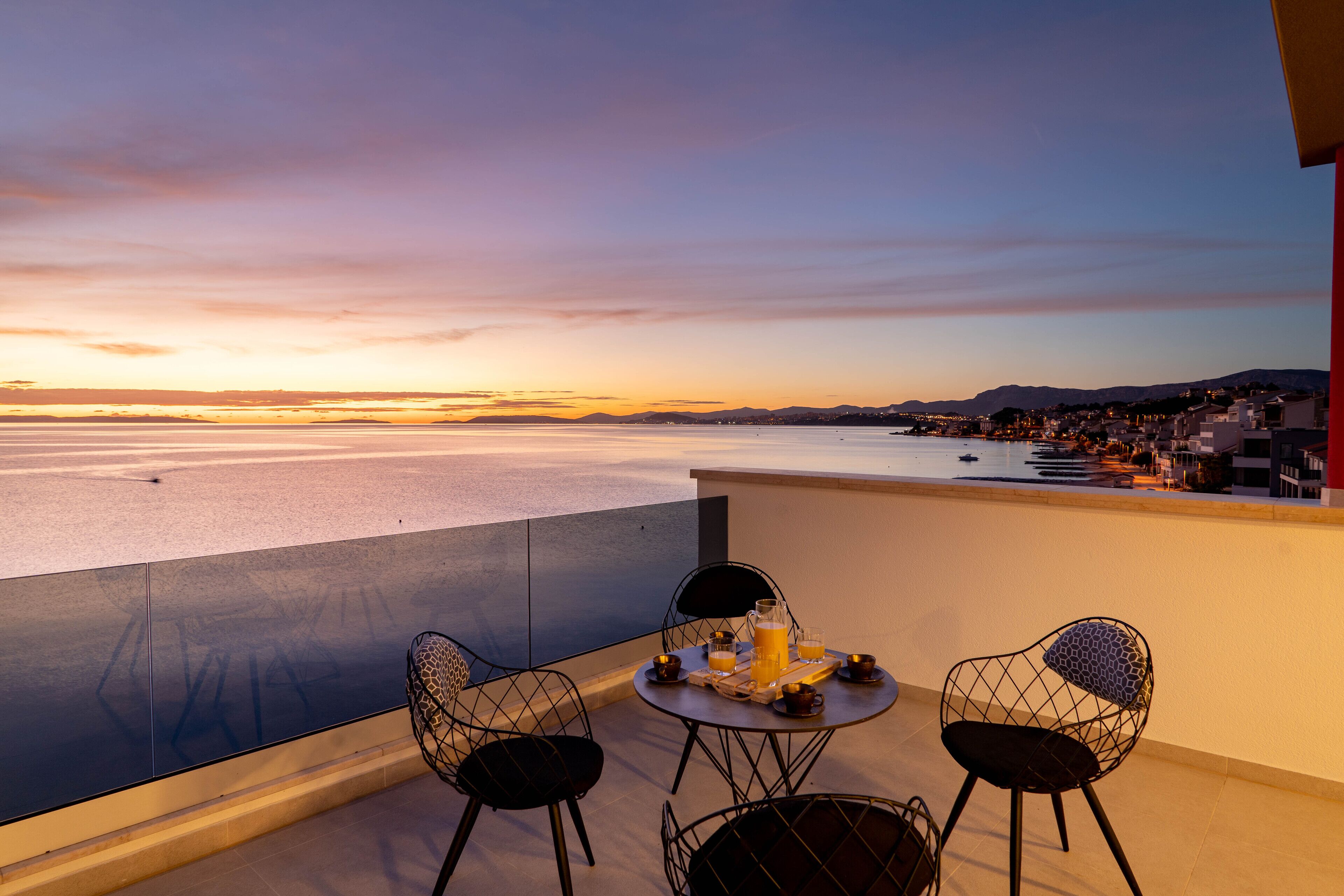 A balcony with a table set for two, overlooking a coastal cityscape during sunset.