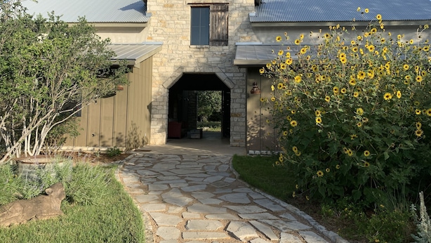 A stone building with a metal roof, a stone pathway leading to an entrance, and a bush with yellow flowers. - Dog-Trot Home on secluded acreage with pool and game room. (Spring Branch)