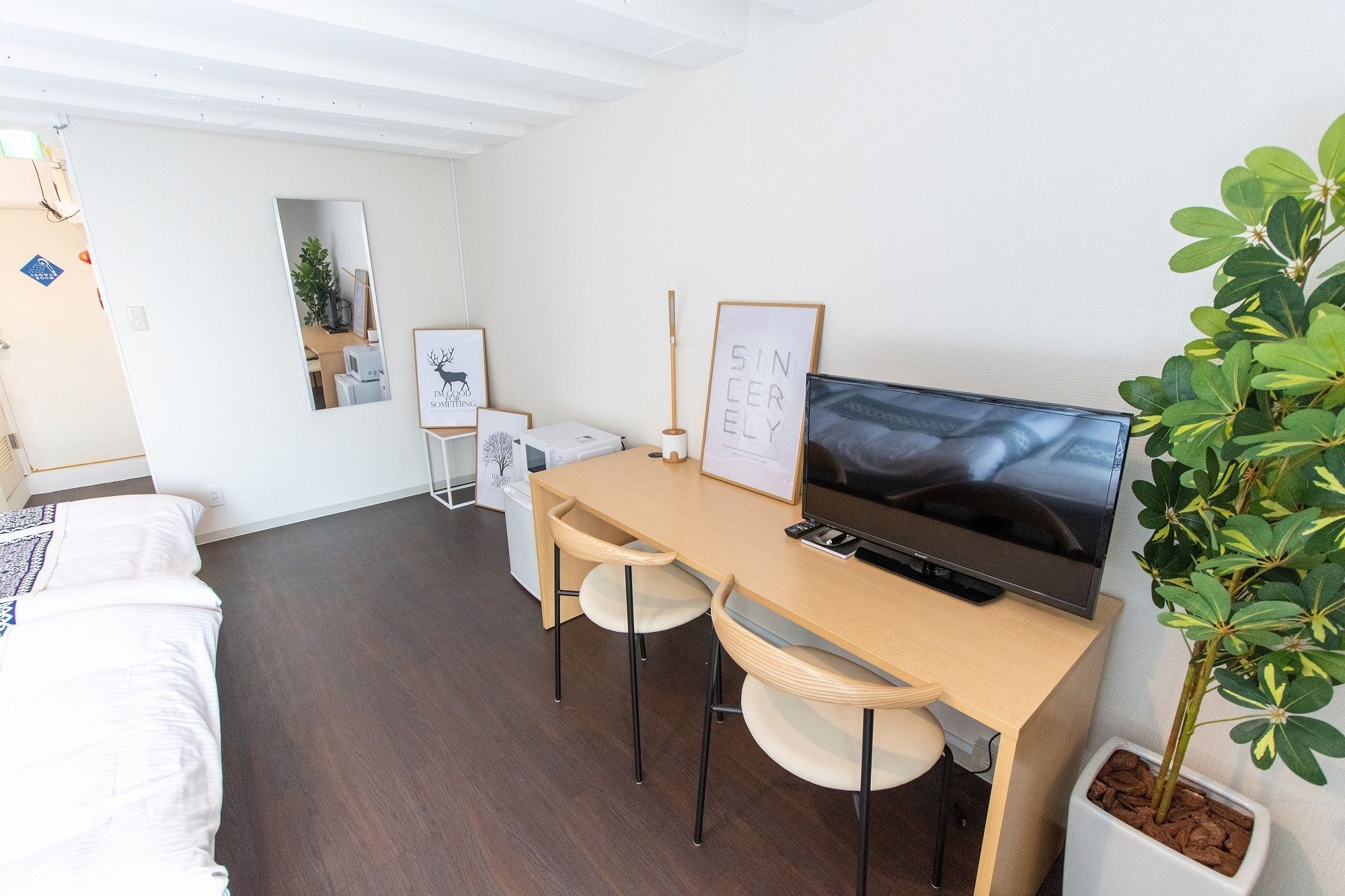Una stanza compatta con una scrivania in legno, due sedie, un letto, una TV e una pianta in vaso.