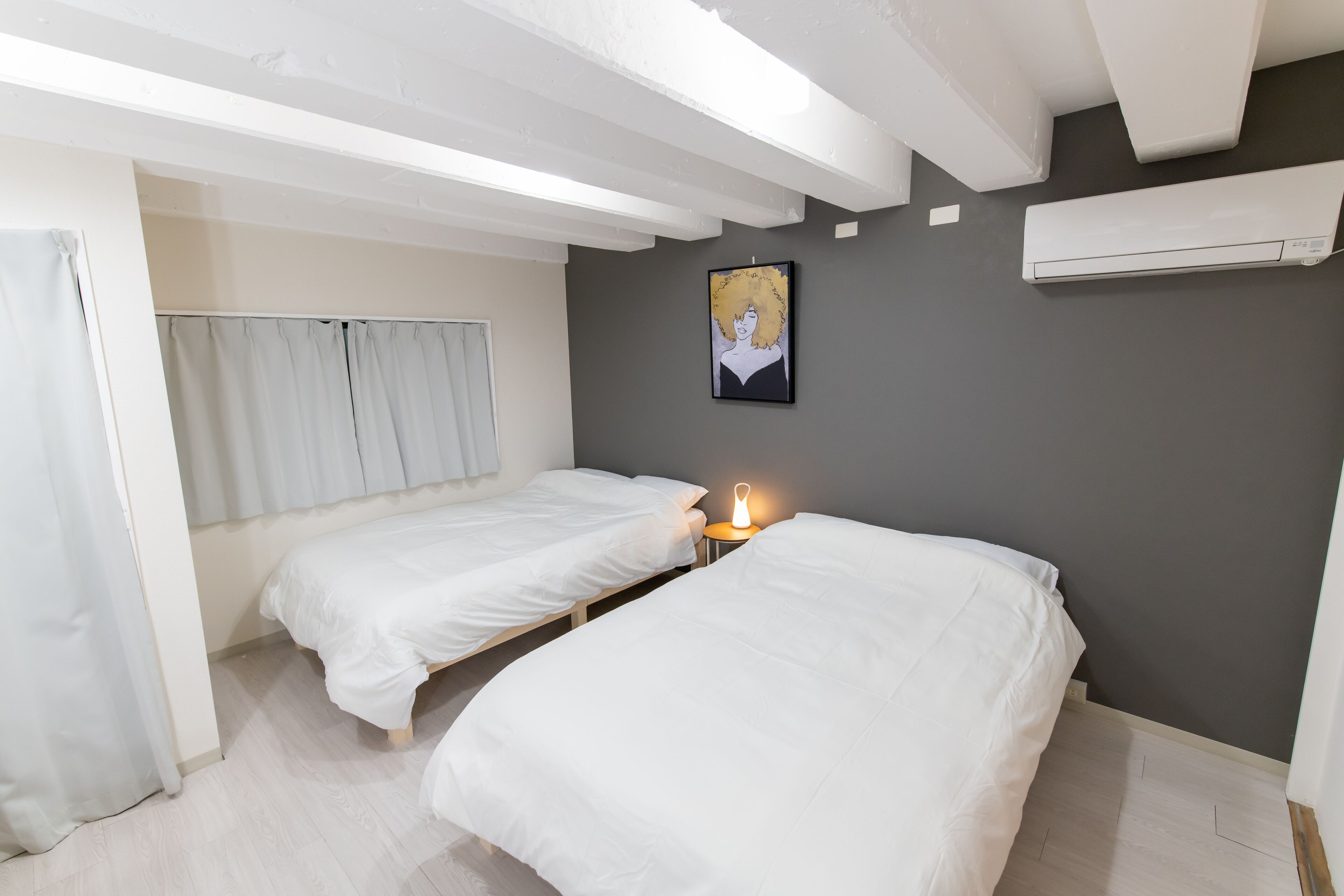 A room with two single beds, a bedside table with a lamp, a wall-mounted air conditioner, and a framed picture on the wall.