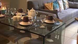 En moderne stue med et glasbord dækket til fire personer, en grå sofa med farverige puder, og et stort vindue med gardiner.